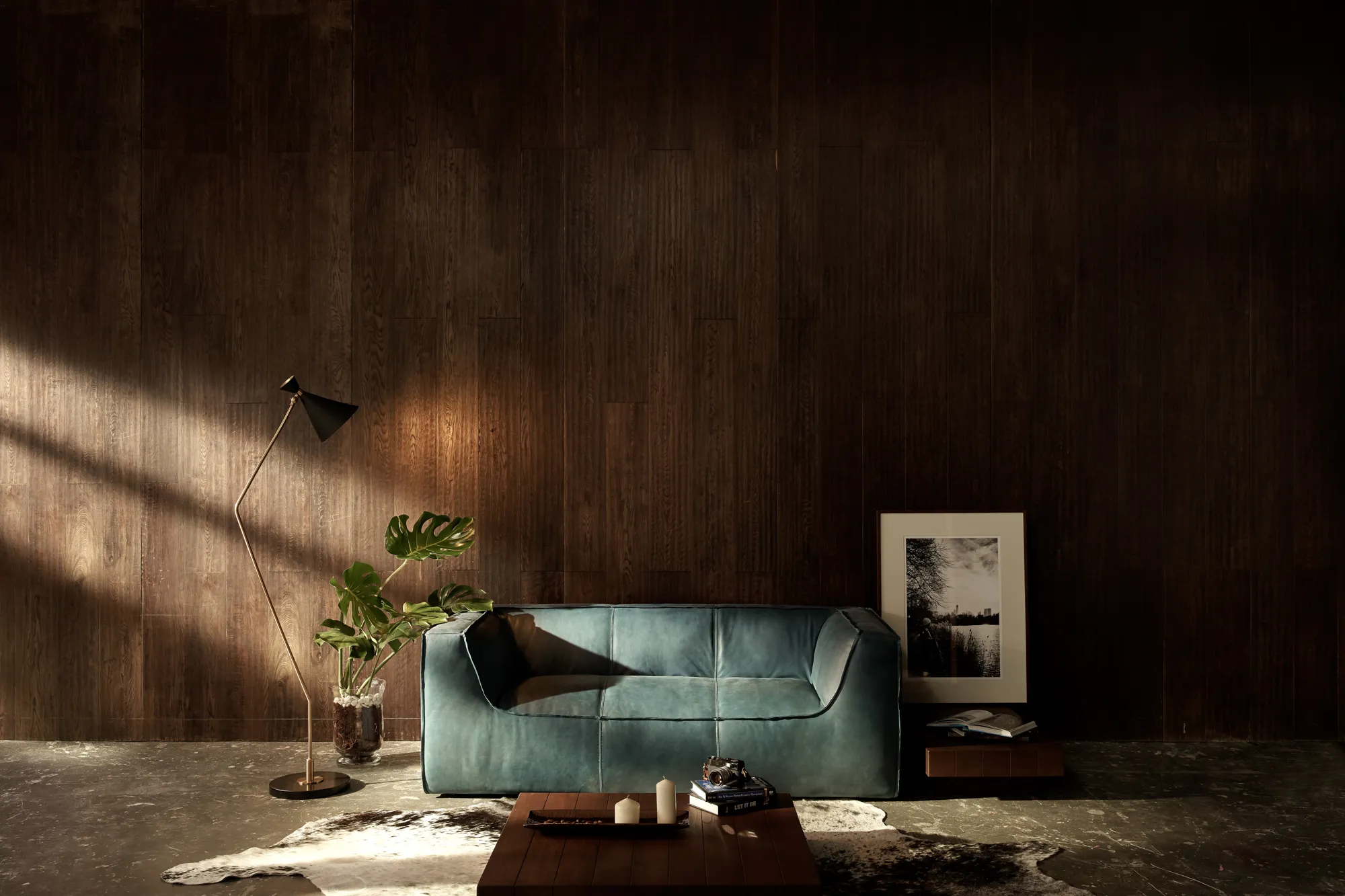 A teal leather sofa and floor lamp against a dark timber wall, lit by a shaft of sunlight