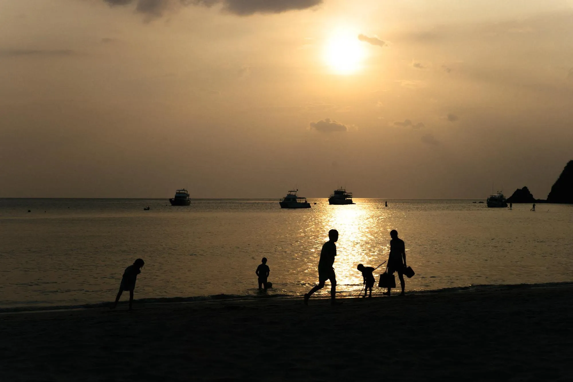 Silhouettes of a family walking along the shore at sunset, boats on the water
