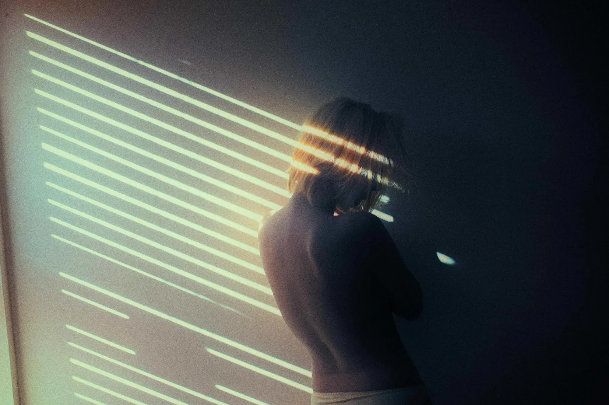 A woman with her back turned, striped light from blinds falling across her and the wall