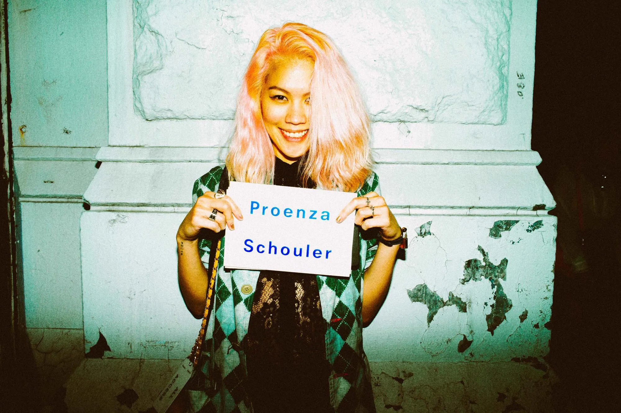 A smiling woman with pink hair holds a sign reading “Proenza Schouler”