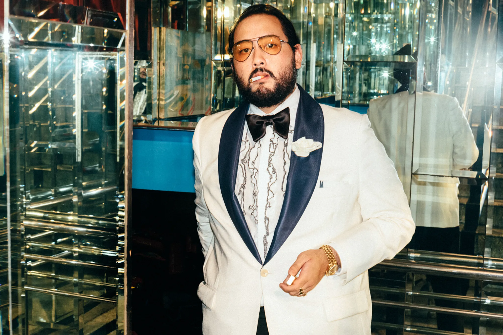 A bearded man in a white tuxedo jacket and amber glasses in a mirrored bar